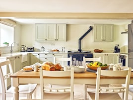 Una cocina con armarios blancos, una mesa de comedor de madera y sillas. Hay una estufa, un fregadero y varios utensilios de cocina en las encimeras.