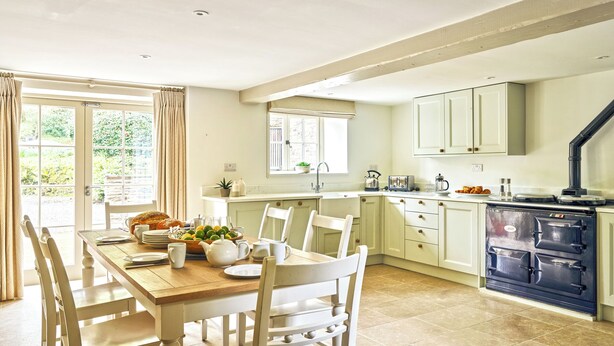 A kitchen with a dining table, chairs, a stove, and a window with curtains. - Gitcombe Coach House (Totnes)