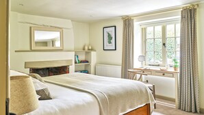 Un dormitorio con cama, mesita de noche, espejo, radiador, ventana con cortinas y una estantería con libros.