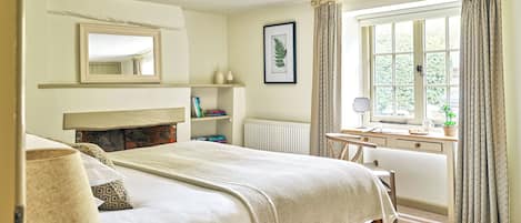 A bedroom with a bed, bedside table, mirror, radiator, window with curtains, and a shelf with books.