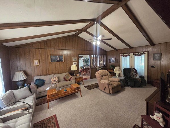 A living room with a high ceiling, wooden walls, a ceiling fan, a sofa, a chair, a coffee table, and a fireplace.