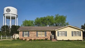A single-story house with a brick facade and a small porch, a water tower in the background, and a well-maintained lawn. - This cozy 3 bed 2 bath house will remind you of a weekend at Grandmother's. (Albertville)