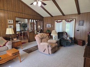 A cozy living room with a wooden paneled wall, a ceiling fan, a sofa, a chair, a small table with a lamp, and a rug.