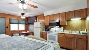 Ein Schlafzimmer mit einem Bett, einem Deckenventilator und einem Fenster. Der Raum verfügt über Holzschränke und eine Küchenzeile mit Mikrowelle, Herd und Kühlschrank.