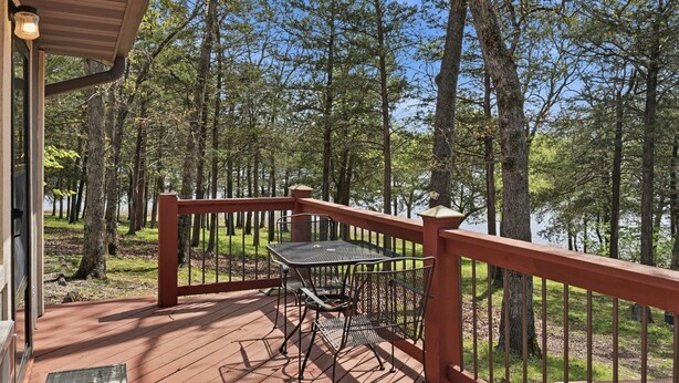 Una terraza de madera con una mesa y sillas de metal, con vista a un área boscosa y un lago.