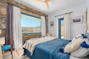 A bedroom with a stone wall, a bed with blue bedding, a wooden chair, and a view of the landscape. - Ikos Kythnos (Kithnos)