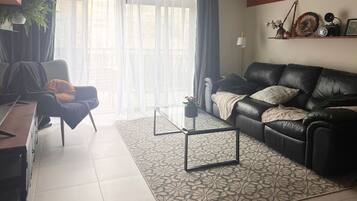 A living room with a black leather sofa, a glass coffee table, a patterned rug, a desk with a chair, and a large window with curtains.