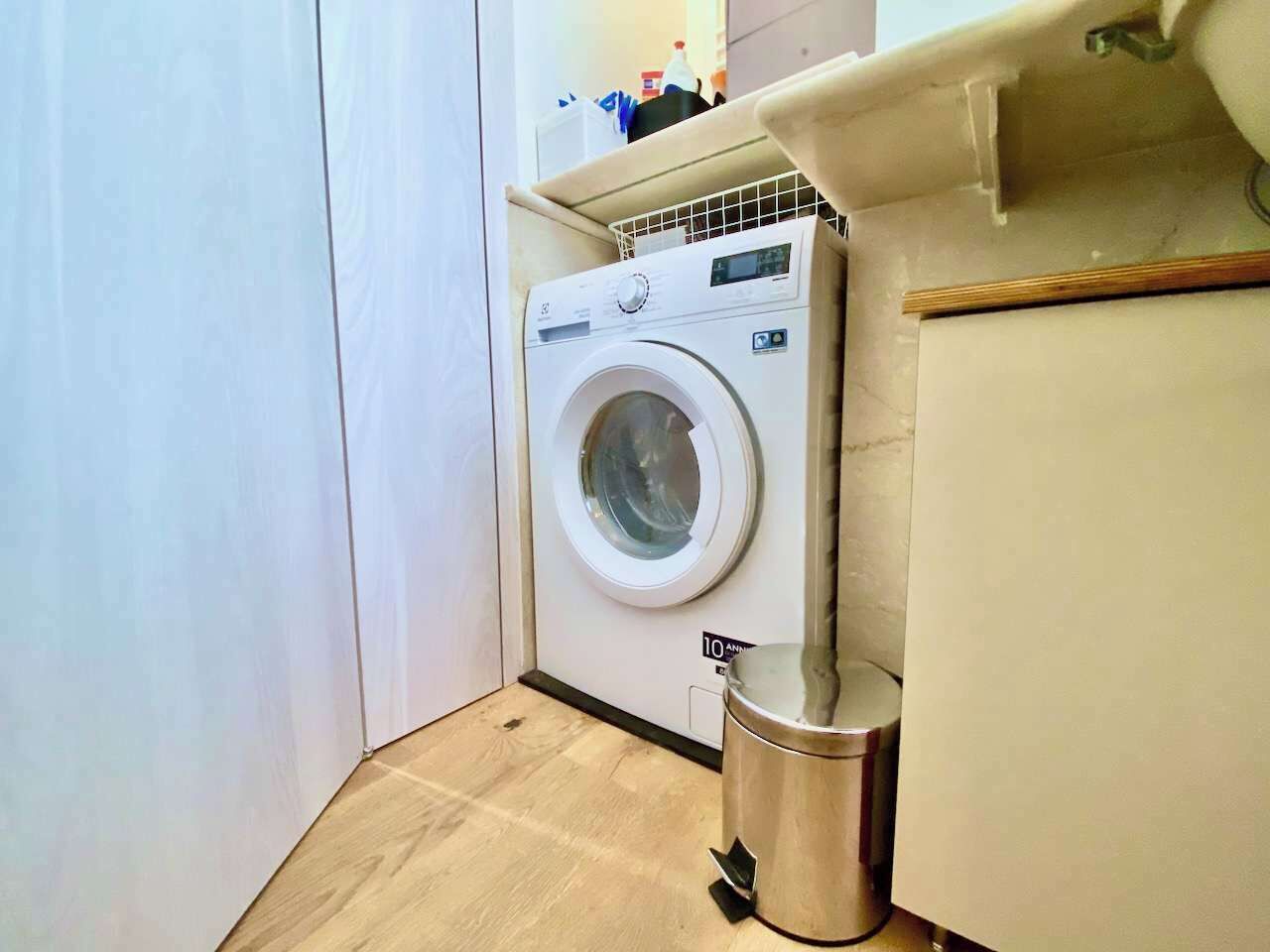 A compact laundry room with a front-loading washing machine, a stainless steel trash can, and a wooden floor. - Luxury 2bed-2bath in top location (Milano)