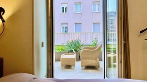 Et hotelværelse med en seng, en lille balkon med fletmøbler og udsigt til en bygning med vinduer.