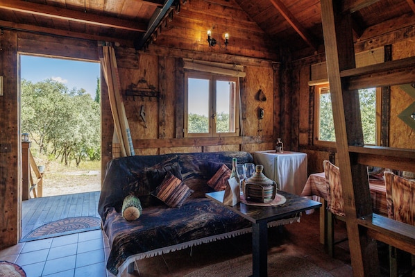 A wooden cabin with a sofa, a table with a bottle and glasses, and a window overlooking trees.