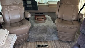 Interior de vehículo recreativo con dos sillones de cuero reclinables, una mesa consola central, un televisor montado y una ventana con cortinas.