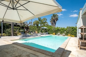 Una piscina amb gandules i un parasol, envoltada de vegetació i palmeres.