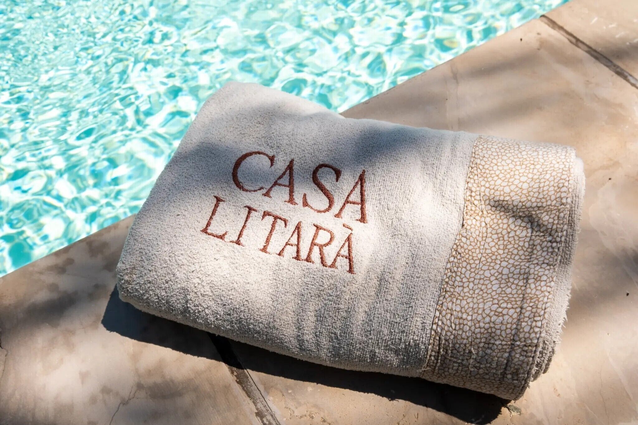 Une serviette ornée d’une broderie « CASA LITARA », posée sur une surface en béton près d’une piscine.
