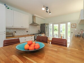 A modern kitchen with white cabinets, a large island with a bowl of fruit, and a dining area with brown chairs. - Millmead House (Weymouth)