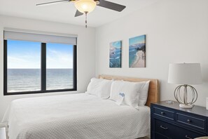 Un dormitorio con un ventanal grande, una cama con ropa de cama blanca, un ventilador de techo y dos cuadros colgados en la pared.