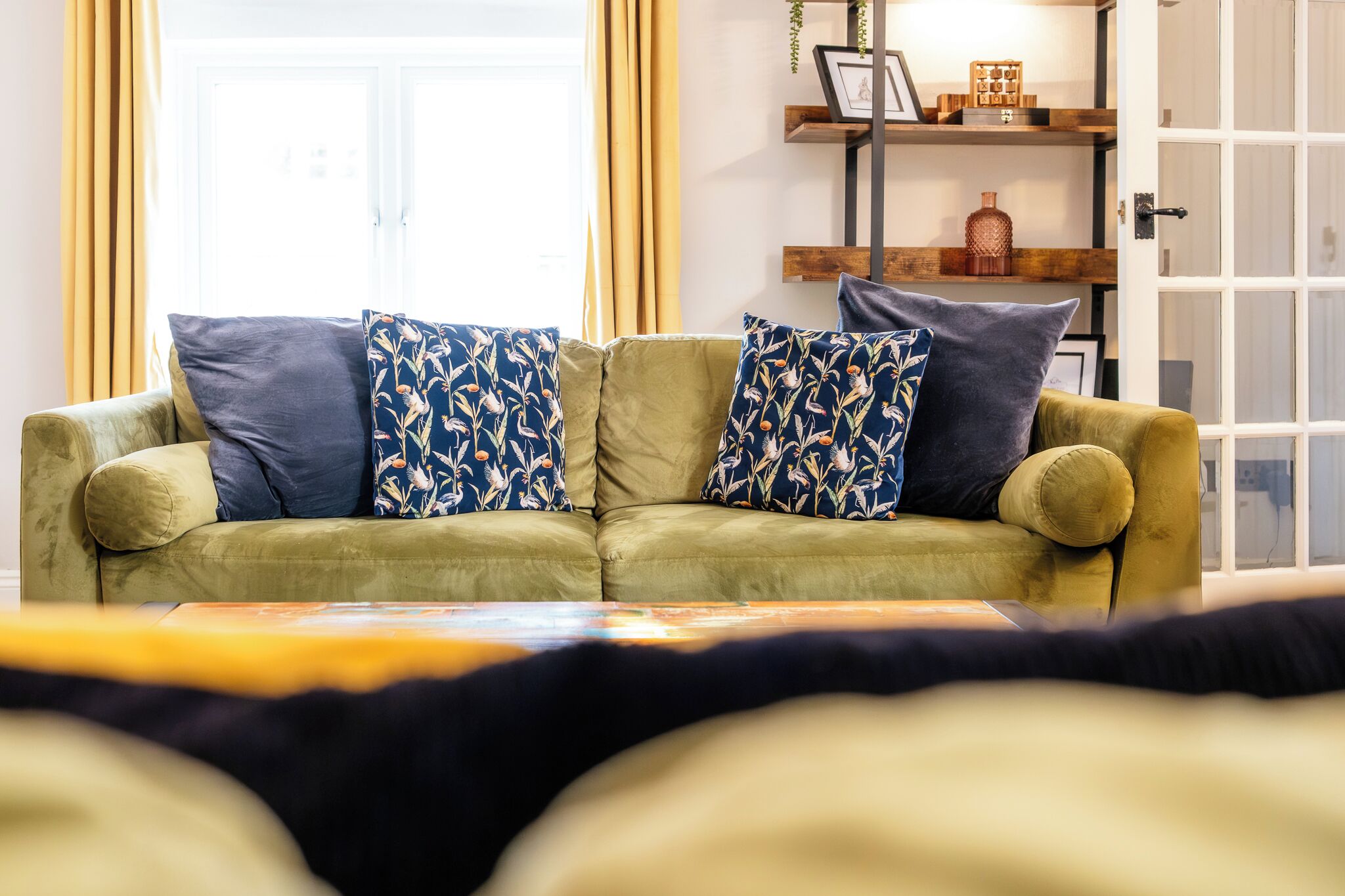 Un sofá verde con almohadones estampados, una estantería de madera con adornos y una puerta blanca con paneles de vidrio.