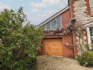 A brick house with a garage door, a stone wall, and a garden with green foliage and purple flowers. - Millmead House Annex (Weymouth)