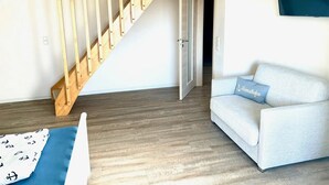 Una habitación con una escalera de madera, un sofá de color claro con un cojín y un televisor de pantalla plana en la pared.