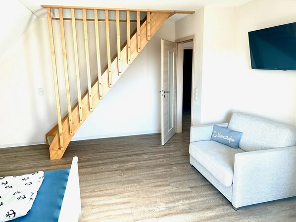 A room with a wooden staircase, a light-colored sofa with a cushion, and a flat-screen TV mounted on the wall.