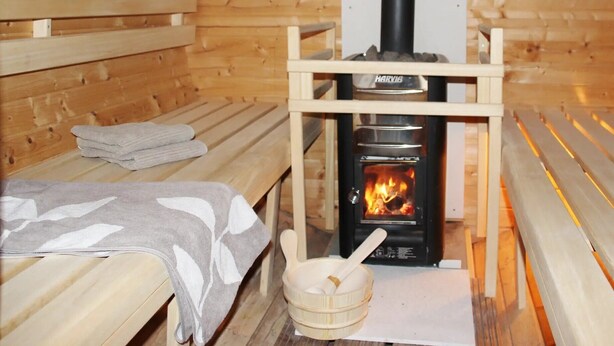 Una sala de sauna con bancos de madera, un banco con una toalla, un cubo de madera y un calentador de sauna con la etiqueta 'HARVIS'.