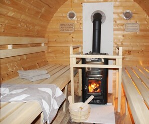 Een sauna met houten banken, een bank met een handdoek, een houten emmer en een sauna kachel met de aanduiding 'HARVIS'.