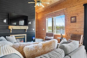 Ein gemütliches Blockhaus-Interieur mit Kamin, bequemen Sitzgelegenheiten, einem Fernseher und einem großen Fenster mit Blick ins Freie.