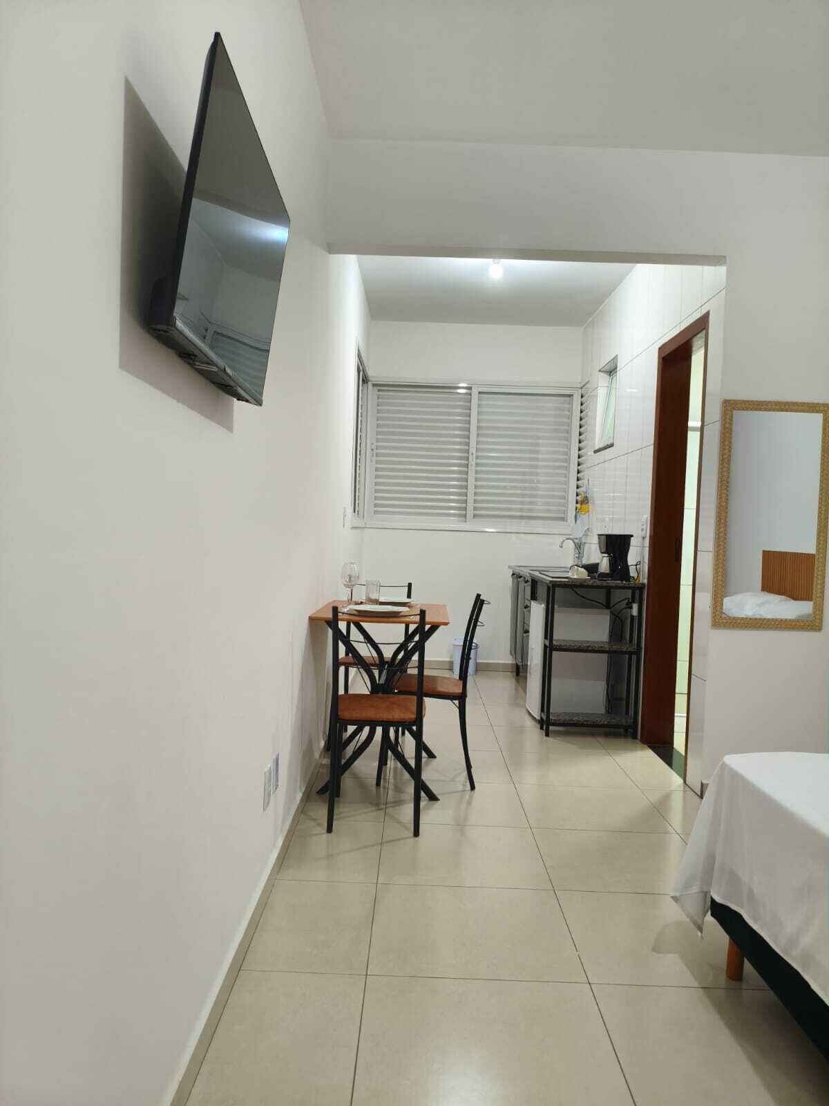 Habitación pequeña con cama, una mesa de comedor con sillas, televisor montado en la pared y ventana con persianas.