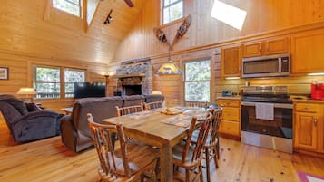 Interior de una cabaña de madera con una mesa de comedor, sillas, una chimenea de piedra, un sofá, un televisor y una zona de cocina con microondas y estufa.