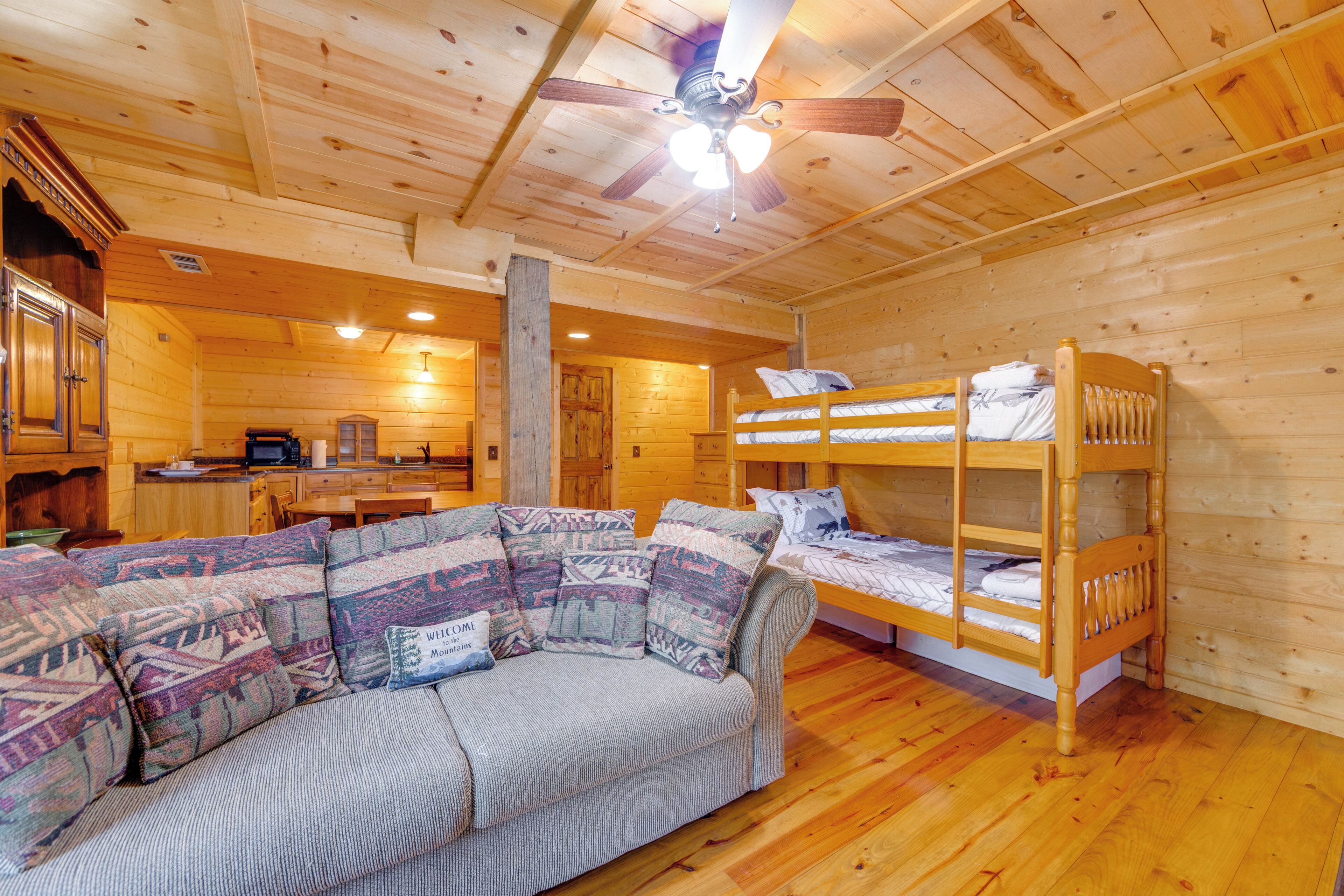 A log cabin interior with a sofa, bunk beds, a kitchen area, and a ceiling fan.