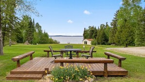 Une terrasse en bois avec une table et des chaises, entourée de pelouse et de fleurs, d’où l’on peut admirer un plan d’eau et des arbres.
