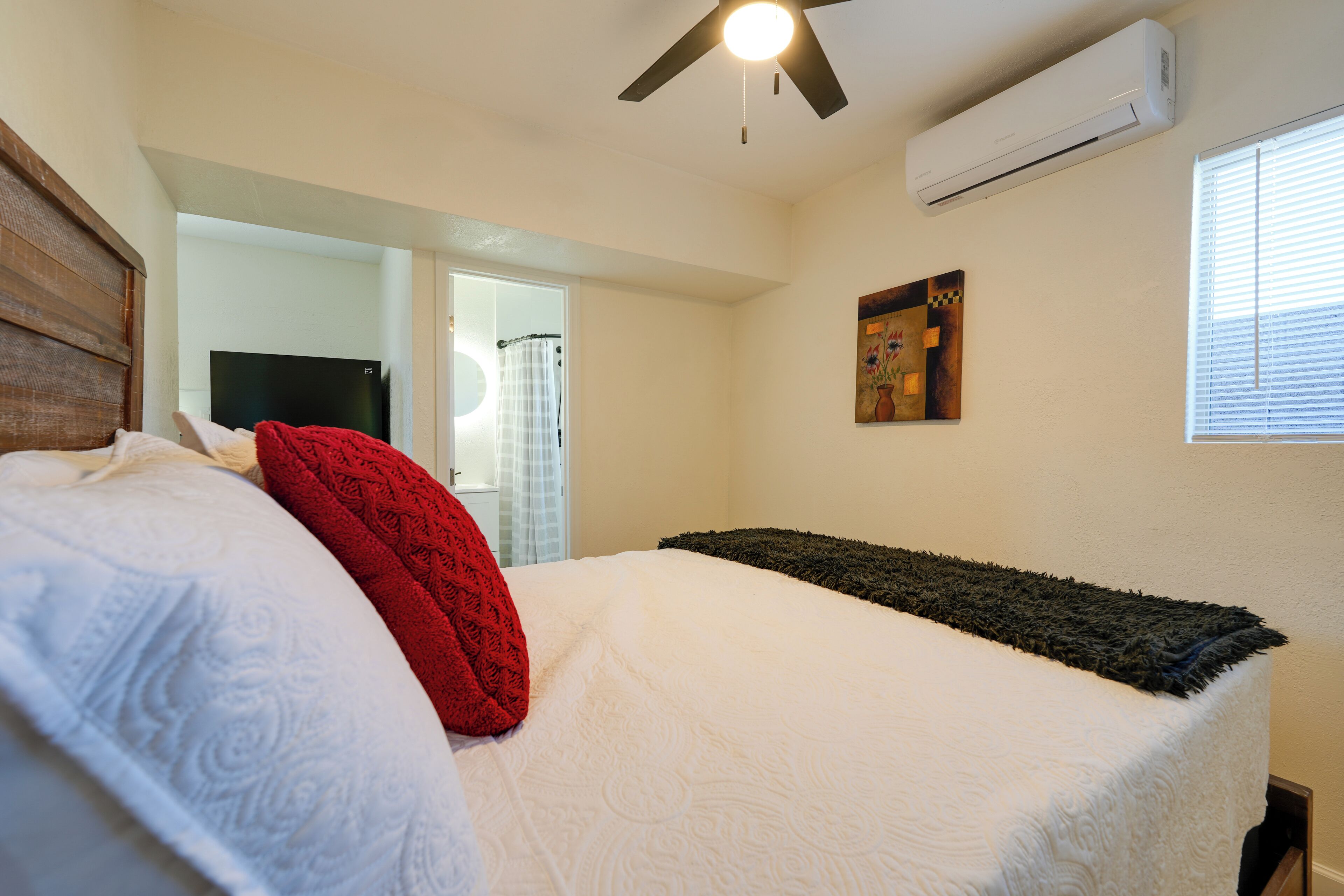 A bedroom with a bed, a ceiling fan, an air conditioner, a window with blinds, and a painting on the wall.