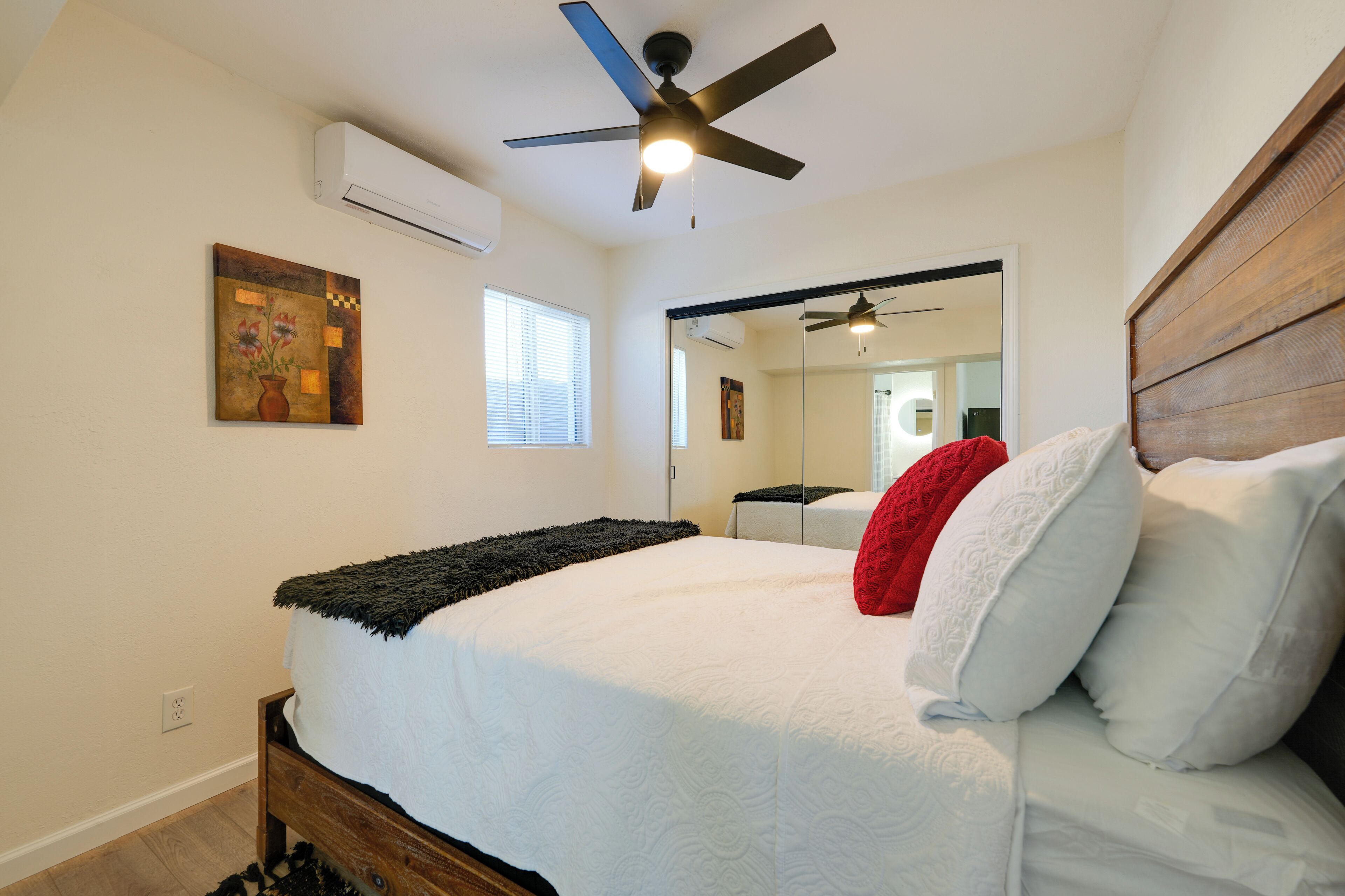 A bedroom with a bed, a ceiling fan, an air conditioner, a window, and a mirror.