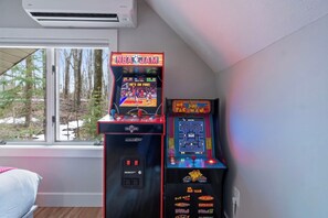 Two arcade machines, one displaying NBA Jam and the other Pac-Man, in a room with a window overlooking trees.