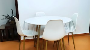 Een ronde tafel met zes witte stoelen, een klein houten bijzettafeltje met een plant, en een effen witte muur.