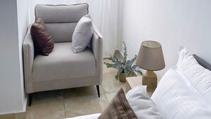 Een kamer met een bed, een fauteuil met kussens, een nachtkastje met lamp en een plant in een pot.