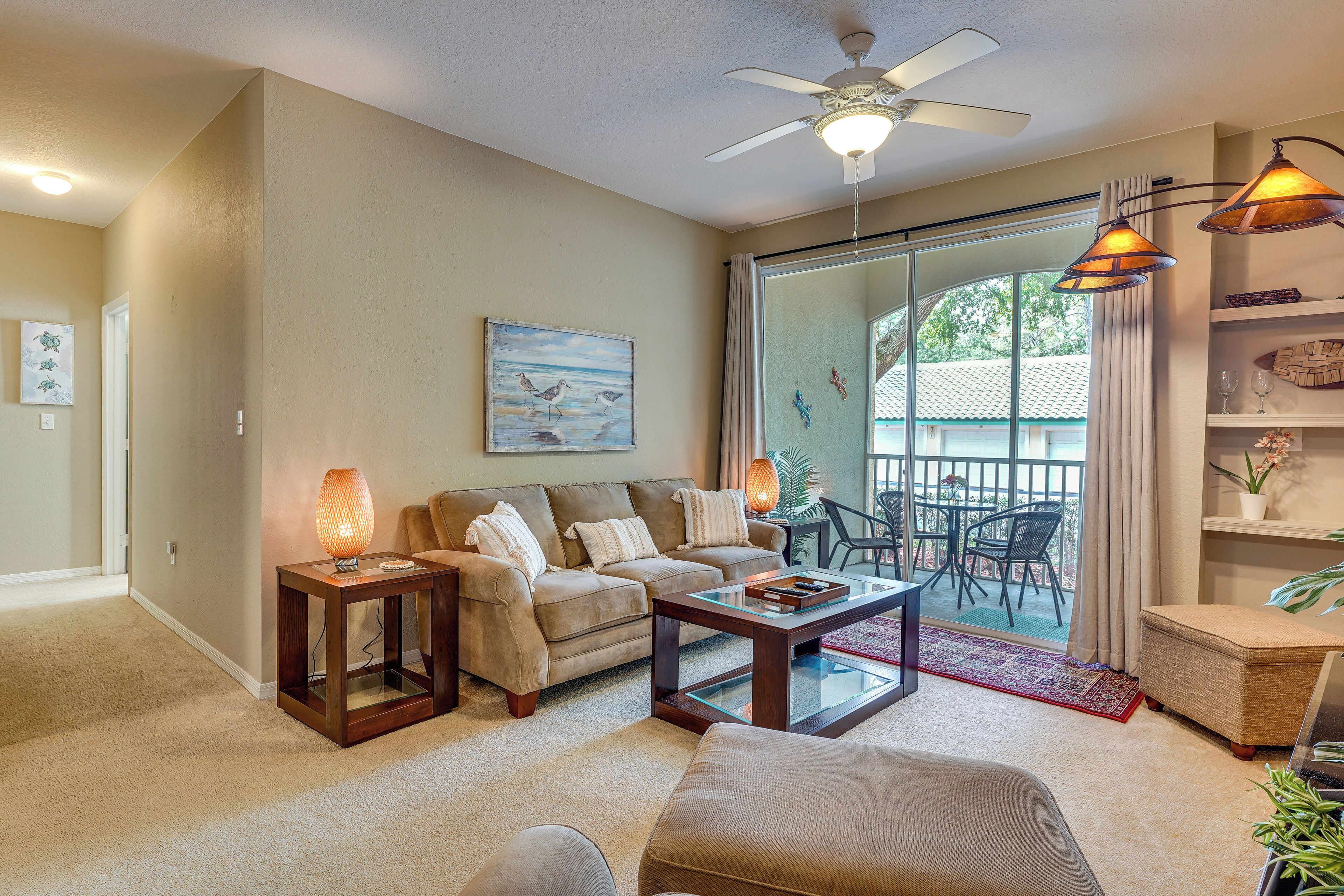 A living room with a sofa, coffee table, and side tables. There is a ceiling fan, a glass door leading outside, and a view of the outdoors.