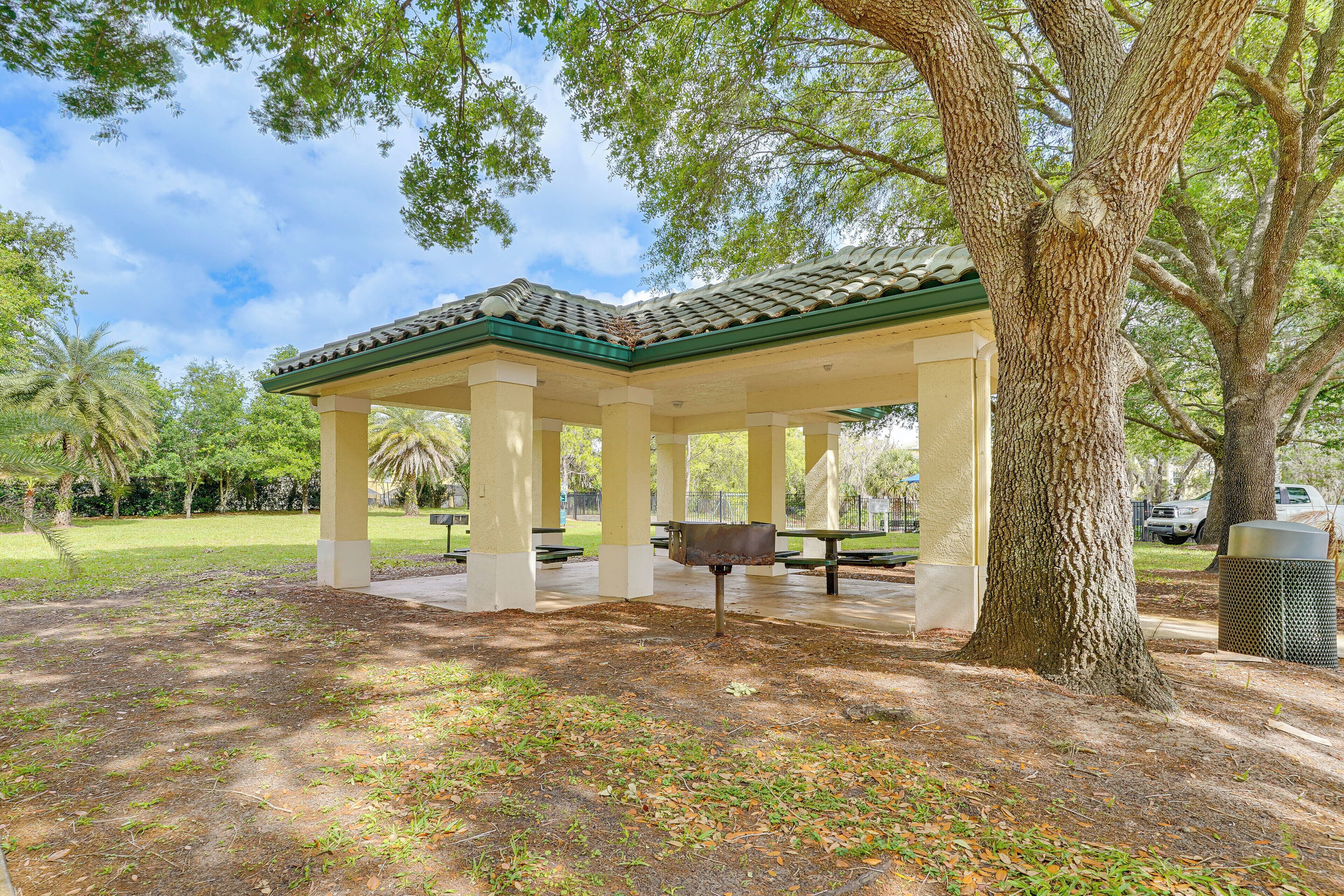 A covered pavilion with a tiled roof, surrounded by trees and a grassy area.