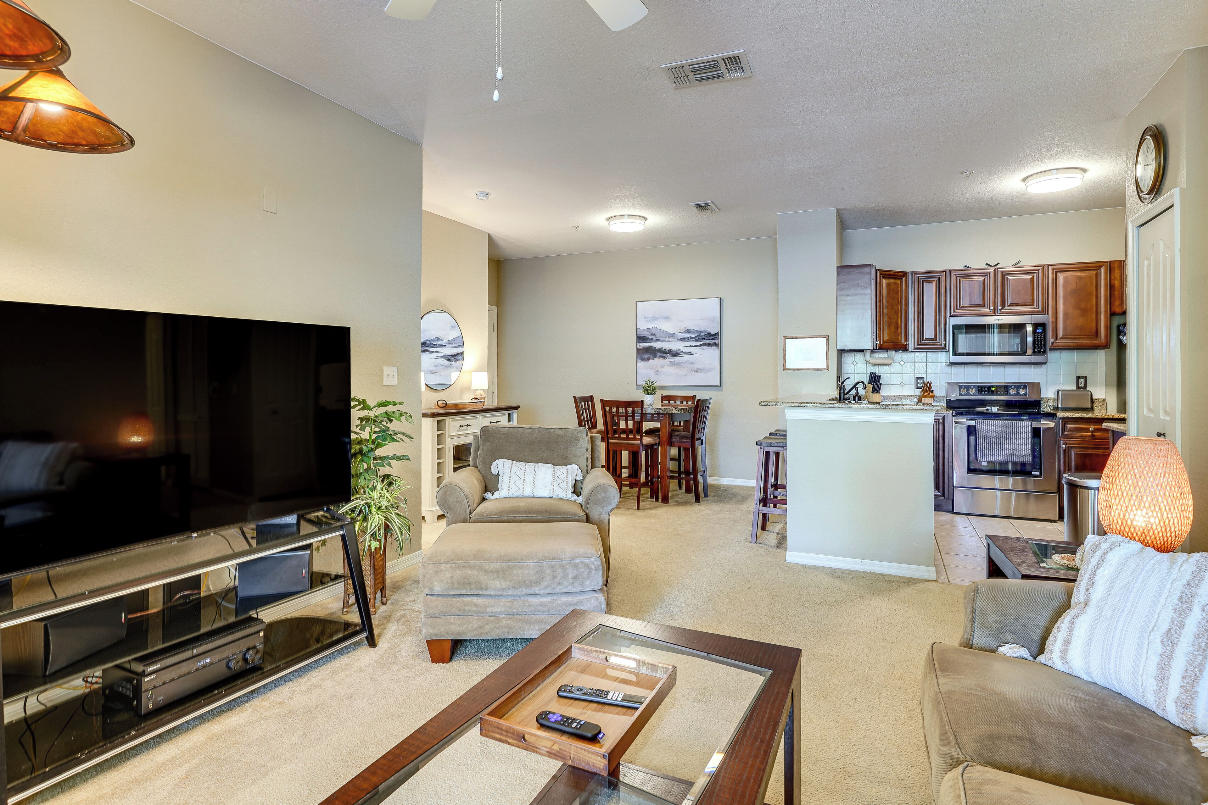 A living room with a flat-screen TV, a coffee table with remotes, a sofa, and a dining area with chairs and a table.