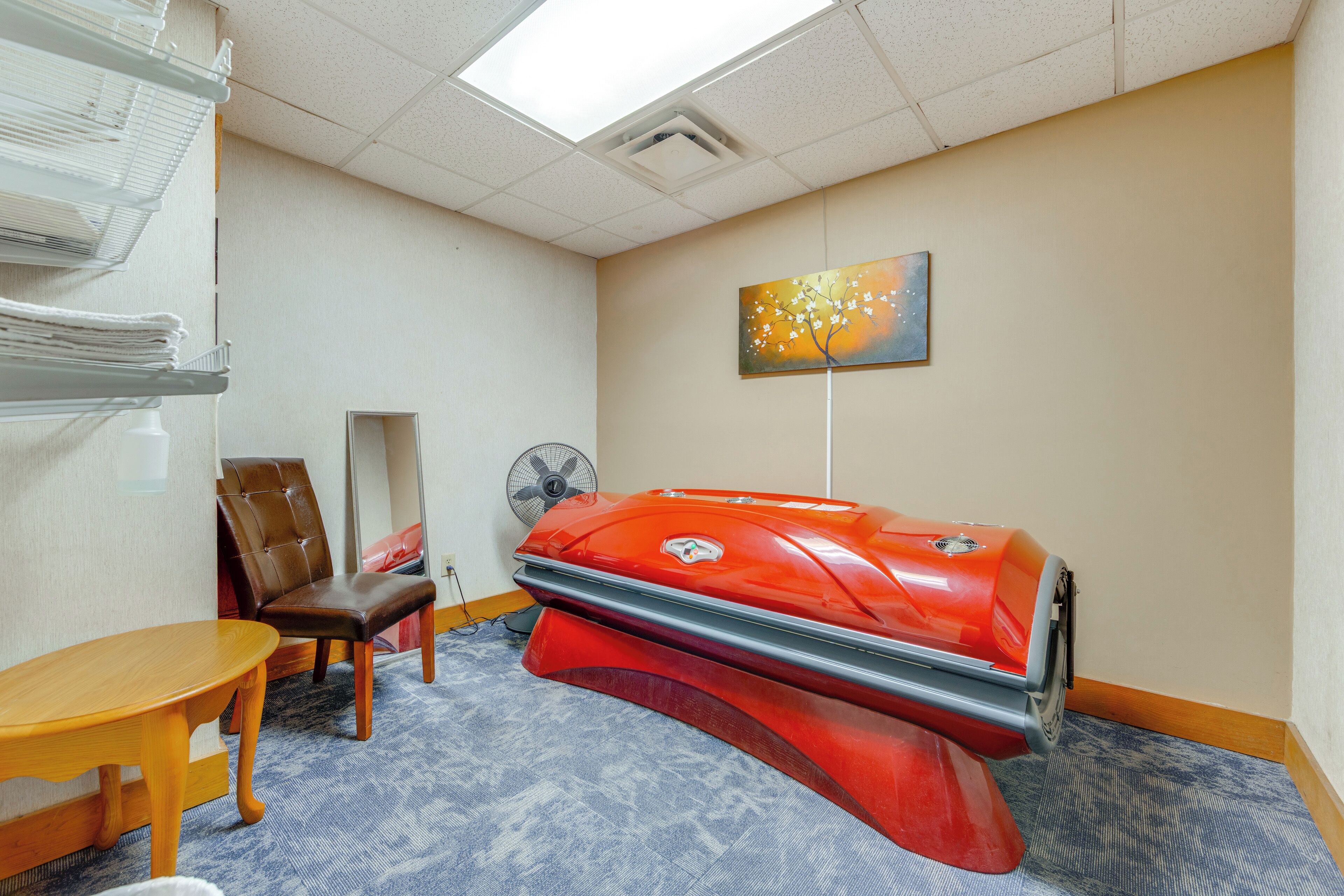 A room with a massage table, a chair, a fan, and a painting on the wall.