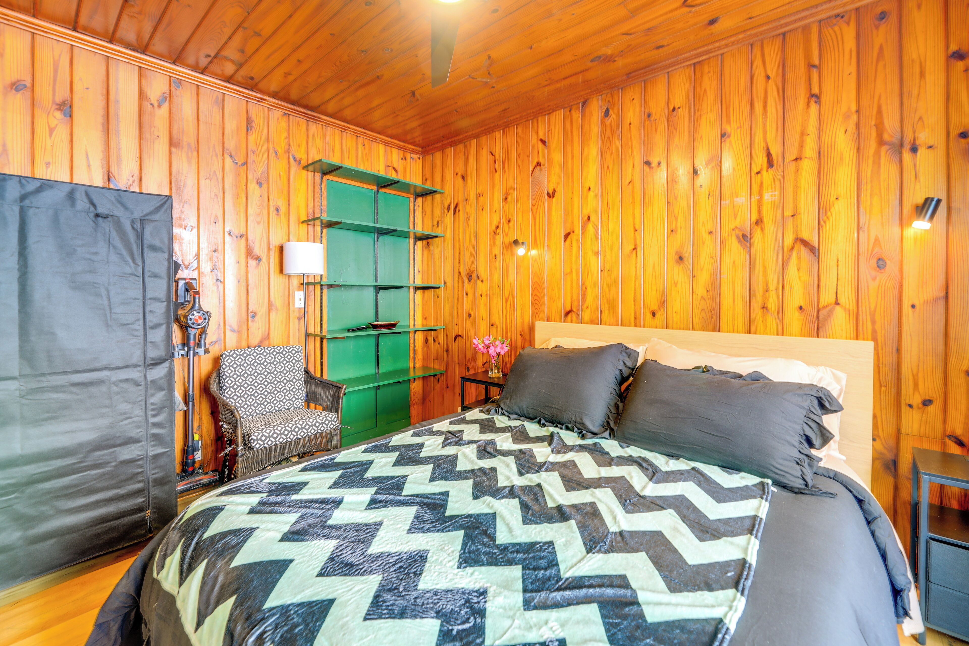 A wooden-paneled room with a bed, a chair, a small table, and a green shelf unit.