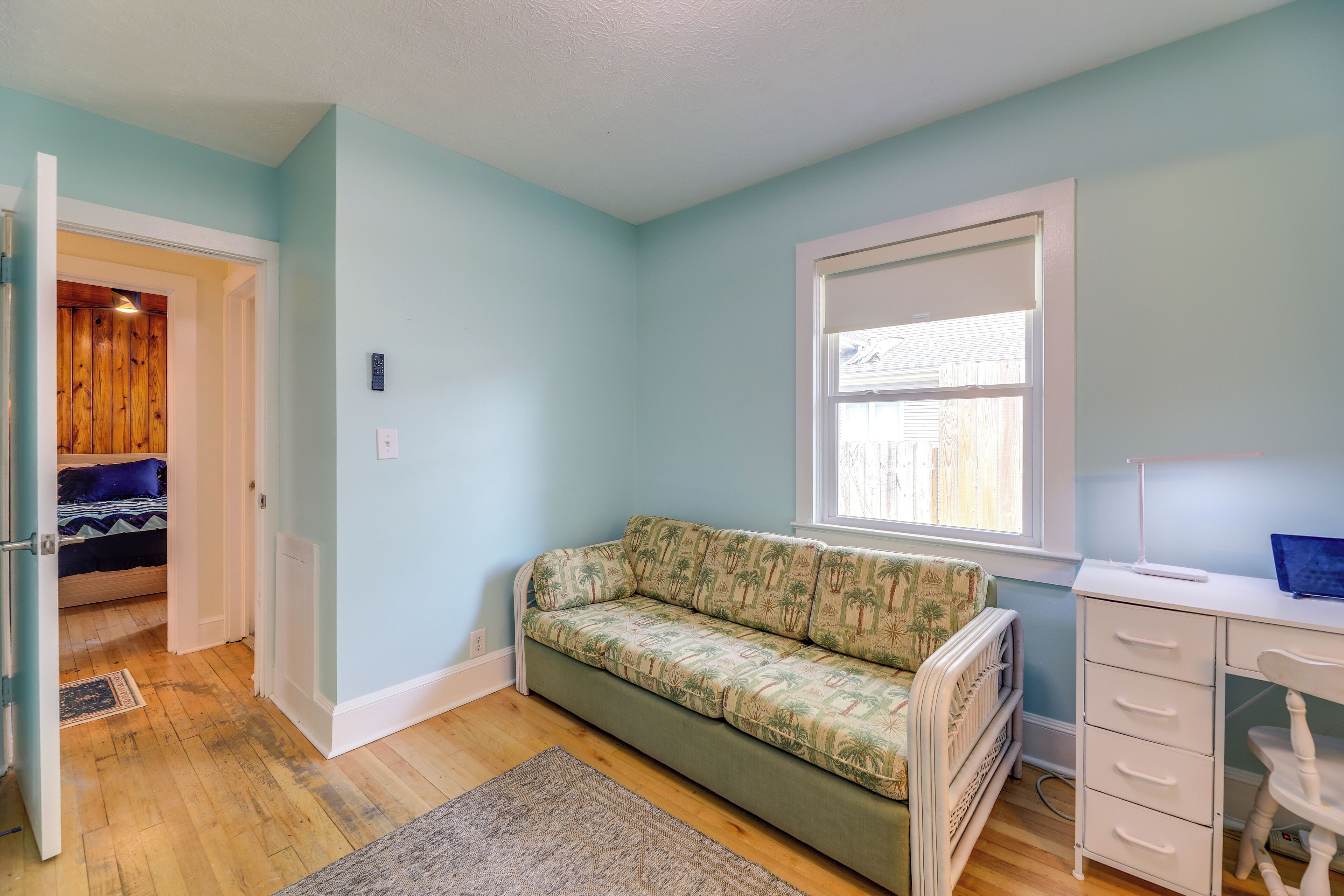 A room with a green sofa, a white desk, a window, and a door leading to another room.