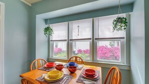 Une table à manger dressée avec des assiettes, des bols et des couverts, entourée de chaises, avec une fenêtre et des plantes suspendues.