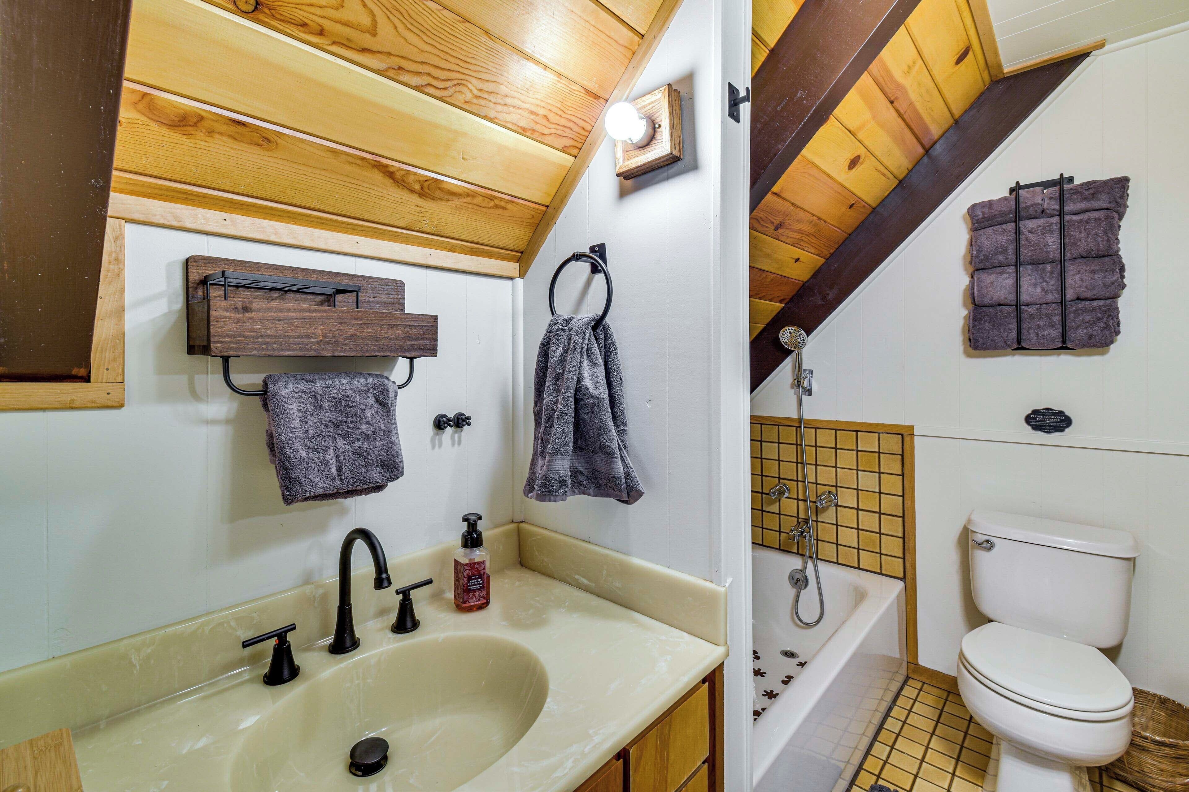 A bathroom with a sink, toilet, and bathtub. There are towels hanging on the wall and a wooden shelf above the sink.