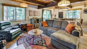 Ein gemütliches Wohnzimmer mit einem großen grauen Sofa, einem Ledersessel und einem Couchtisch. Zwei große Fenster bieten Blick auf Bäume, und die Zimmerdecke ist mit Holzbalken versehen.