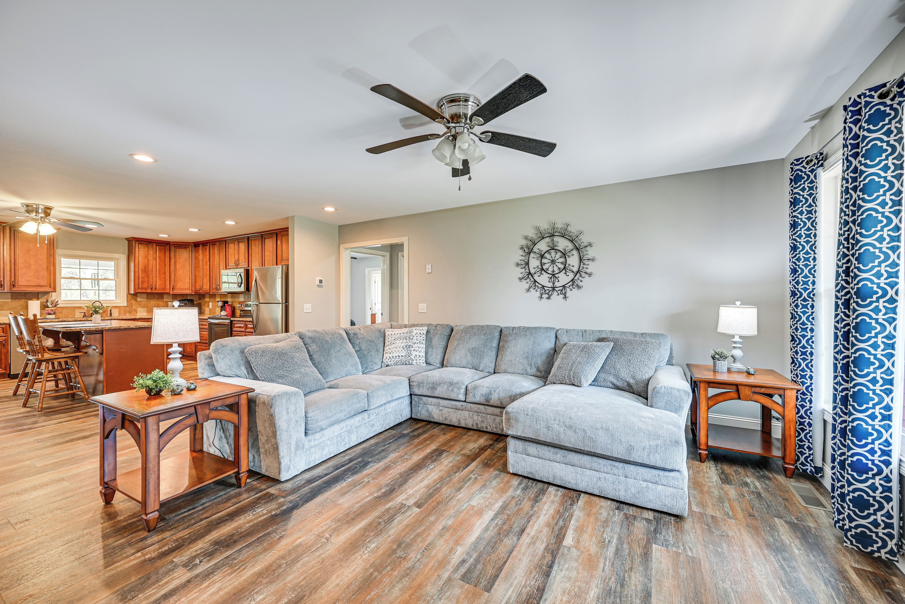 A living room with a sectional sofa, wooden flooring, ceiling fan, and kitchen in the background.