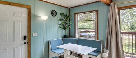 A room with a white door, a bench, a table, a chair, a lamp, a clock, and a window with a view of the outdoors.