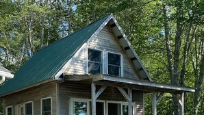 Une petite cabane à un seul étage, avec un toit vert, une véranda dotée d’un banc et une allée en terre.
