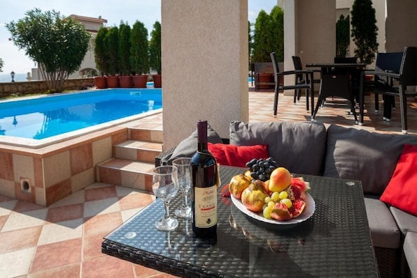A poolside area with a table, a bottle of wine, and a plate of fruit.