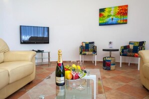 Ein Wohnzimmer mit einer Couch, einem Glastisch, einer Champagnerflasche, Obst und einem Fernseher an der Wand.