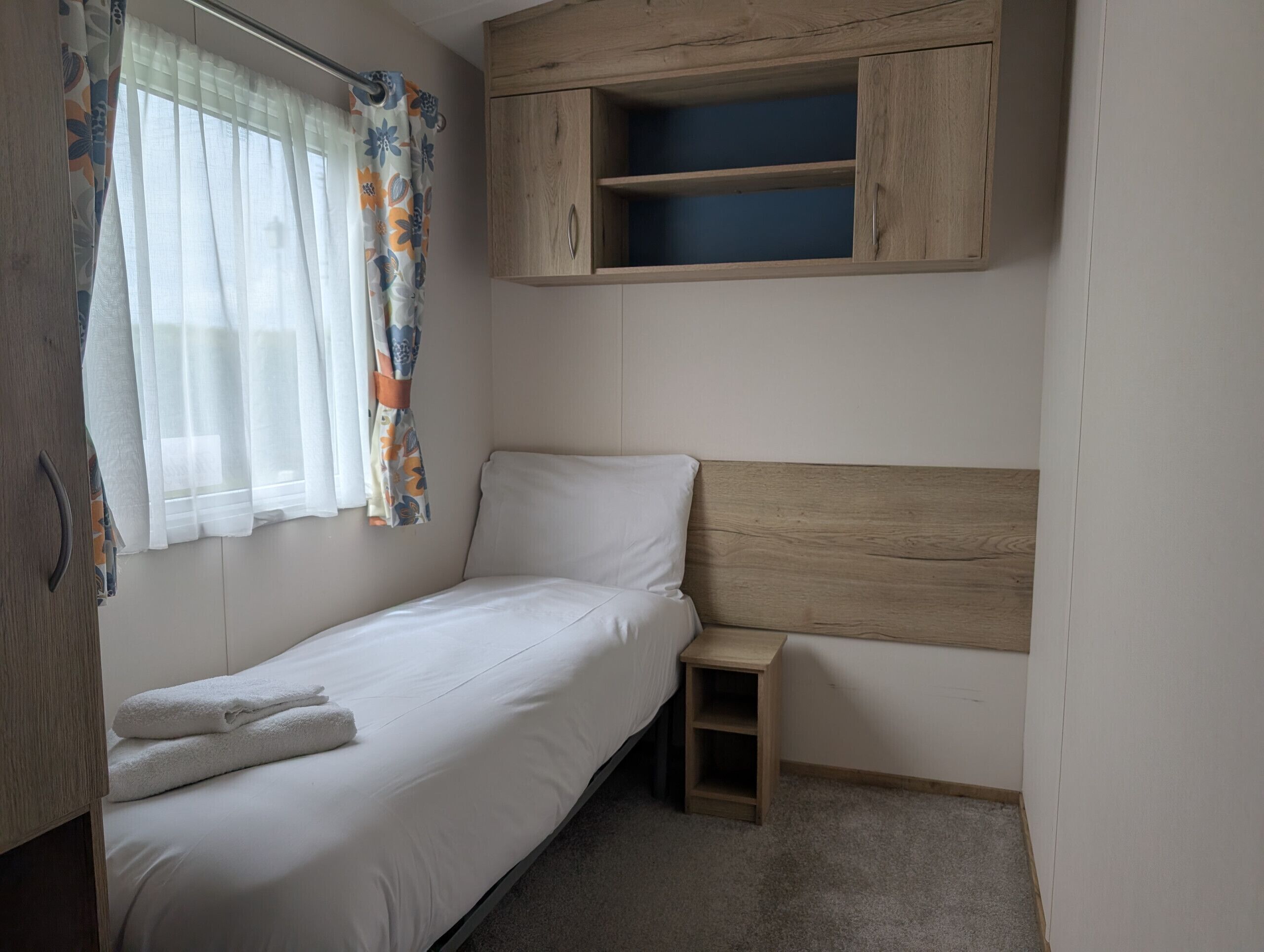 Un lit simple avec une tête de lit en bois, une petite table de chevet en bois, une fenêtre avec des rideaux légers et une étagère intégrée au-dessus du lit.